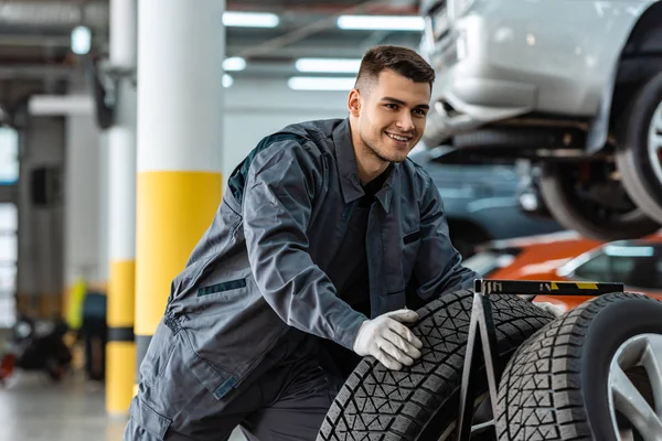 Bello, meccanico sorridente che tiene nuovo pneumatico in officina — Foto stock