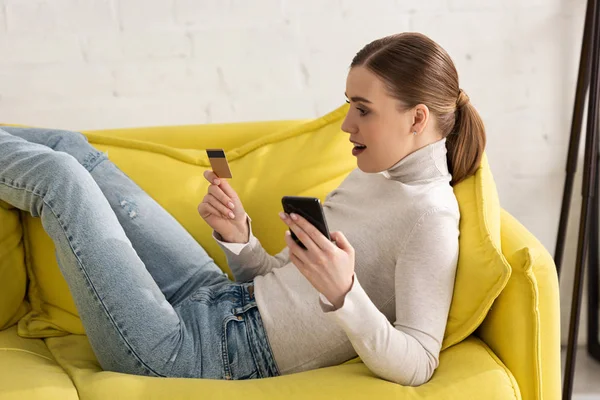 Side view of woman holding credit card and smartphone on sofa at home — Stock Photo