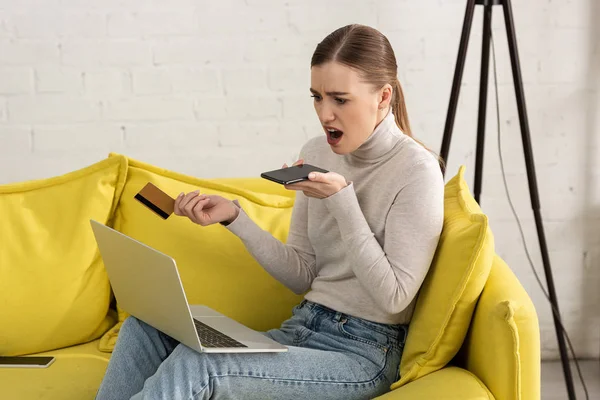 Mulher chocada segurando cartão de crédito e smartphone enquanto olha para laptop — Fotografia de Stock