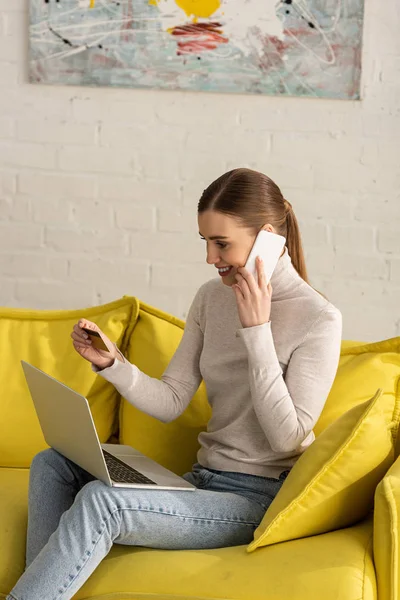 Chica sonriente hablando en el teléfono inteligente mientras sostiene la tarjeta de crédito y el ordenador portátil en el sofá - foto de stock