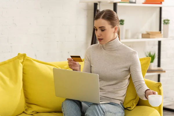 Muchacha atractiva sosteniendo la tarjeta de crédito y la taza de café mientras usa el ordenador portátil en el sofá - foto de stock