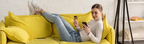 Photo panoramique d'une femme souriante tenant une carte de crédit et utilisant un smartphone sur un canapé à la maison — Photo de stock