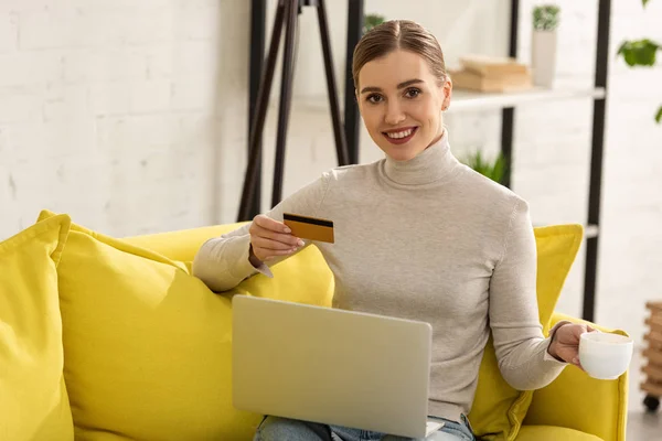 Mulher sorrindo segurando cartão de crédito, laptop e xícara de café no sofá em casa — Fotografia de Stock