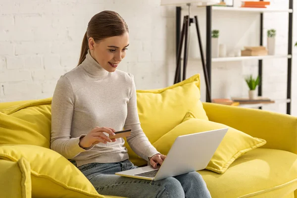 Attraente donna sorridente in possesso di carta di credito e utilizzando il computer portatile sul divano — Foto stock