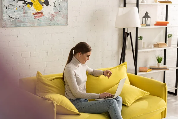 Side view of smiling girl holding credit card and using laptop on sofa in living room — Stock Photo