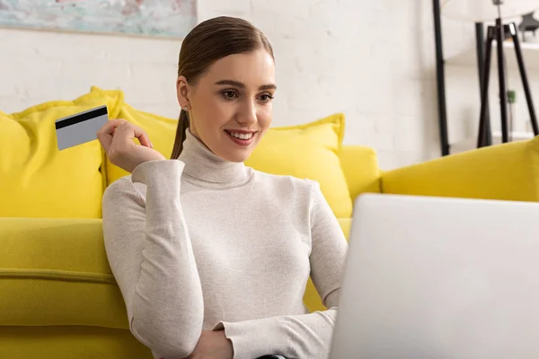 Hermosa chica sonriente con tarjeta de crédito y mirando a la computadora portátil en la sala de estar - foto de stock