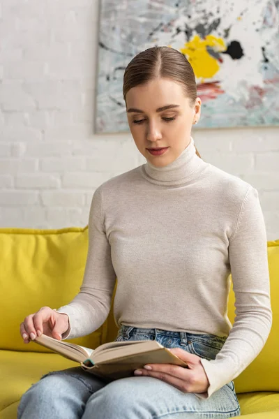 Jolie jeune femme lisant le livre sur le canapé à la maison — Photo de stock