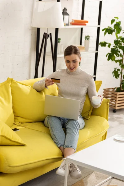 Souriante fille avec ordinateur portable et carte de crédit boire du café sur le canapé dans le salon — Photo de stock