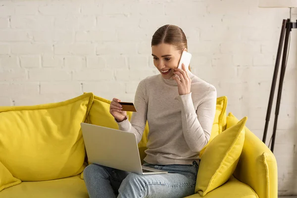 Smiling girl talking on smartphone and holding laptop and credit card on sofa — Stock Photo