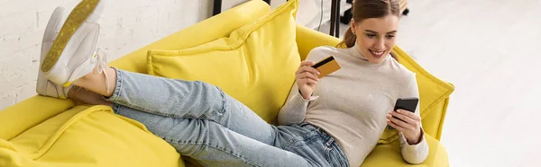 Tiro panorâmico de bela menina sorridente usando smartphone e segurando cartão de crédito no sofá — Fotografia de Stock