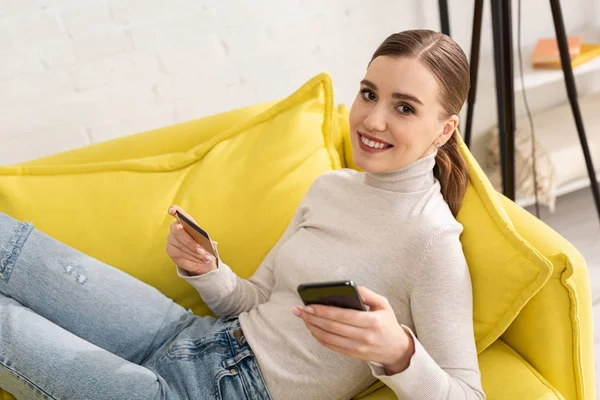 Jolie jeune femme avec carte de crédit et smartphone souriant à la caméra sur le canapé à la maison — Photo de stock