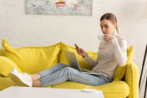 Mujer joven bebiendo café mientras sostiene la tarjeta de crédito y el uso de ordenador portátil en el sofá - foto de stock