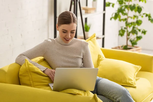 Freelancer sorridente bonita usando laptop no sofá em casa — Fotografia de Stock