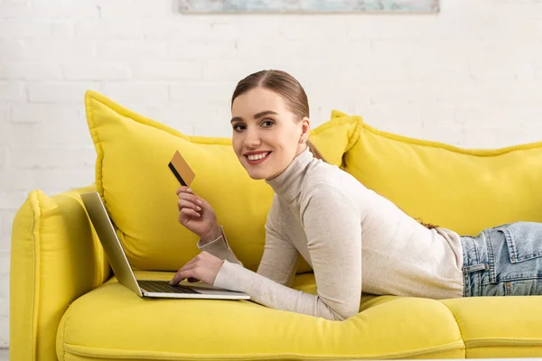 Girl smiling at camera while holding credit card and using laptop on sofa — Stock Photo