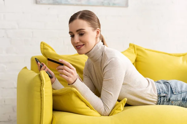 Menina sorrindo olhando para cartão de crédito e segurando smartphone no sofá em casa — Fotografia de Stock