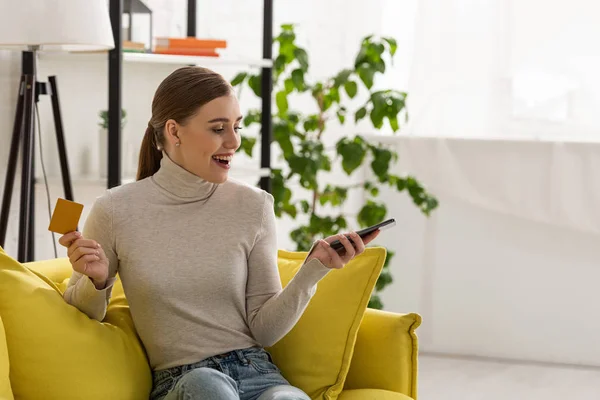 Excited young woman holding credit card and smartphone on sofa at home — Stock Photo