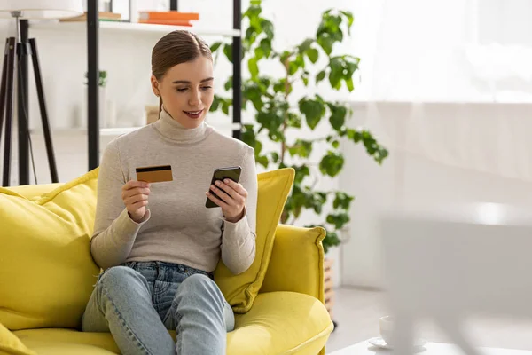 Chica sonriente sosteniendo tarjeta de crédito y el uso de teléfono inteligente en el sofá en la sala de estar - foto de stock