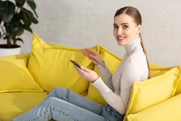 Hermosa mujer sonriendo a la cámara mientras usa el teléfono inteligente y sostiene la tarjeta de crédito en el sofá - foto de stock