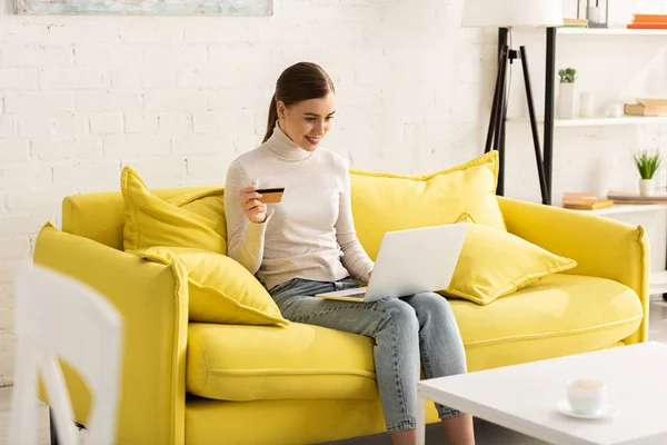 Smiling woman holding credit card and using laptop on couch in living room — Stock Photo