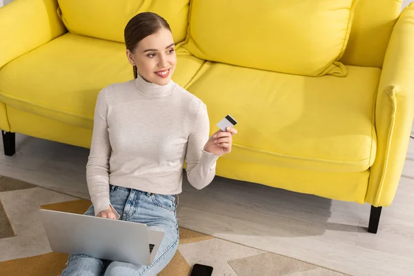 Menina sorridente segurando cartão de crédito e usando laptop no chão na sala de estar — Fotografia de Stock