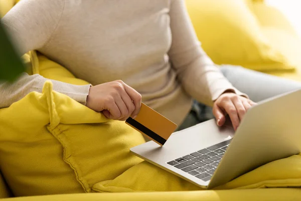 Vista cortada da mulher usando laptop e segurando cartão de crédito no sofá — Fotografia de Stock