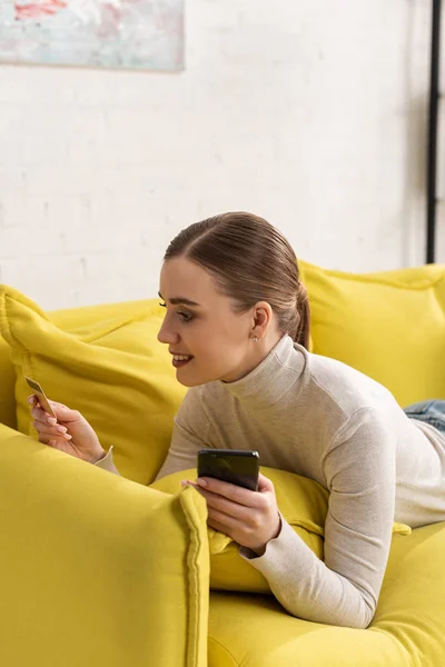 Sorrindo menina atraente segurando cartão de crédito e smartphone no sofá — Fotografia de Stock