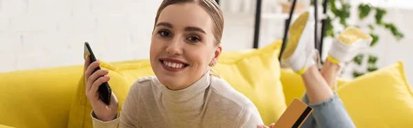 Panoramic shot of woman holding smartphone and credit card and smiling at camera on sofa — Stock Photo