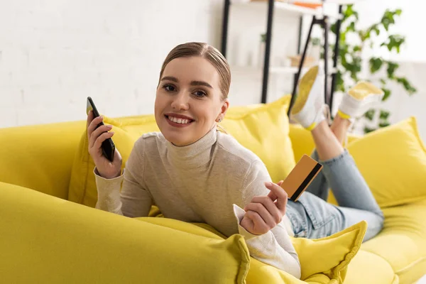 Mujer sonriente sosteniendo teléfono inteligente y tarjeta de crédito mientras está acostada en el sofá - foto de stock