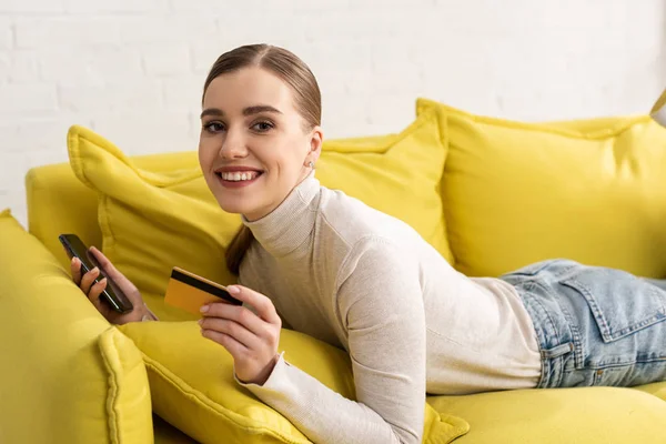Chica sonriente mirando a la cámara mientras sostiene la tarjeta de crédito y el teléfono inteligente en el sofá en casa - foto de stock