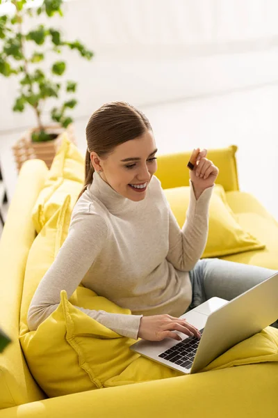 Bella ragazza sorridente usando il computer portatile e tenendo la carta di credito sul divano in soggiorno — Foto stock