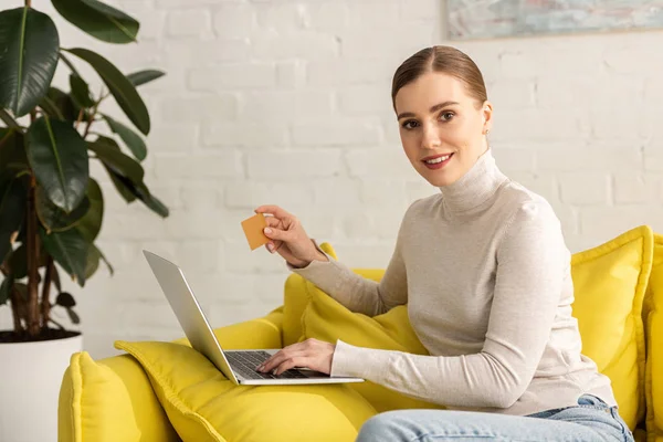 Donna attraente sorridente alla fotocamera durante l'utilizzo di laptop e carta di credito sul divano in soggiorno — Foto stock