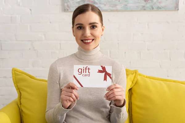 Attraktive junge Frau zeigt Geschenkkarte und lächelt auf Couch in die Kamera — Stockfoto