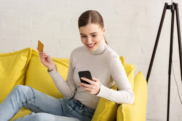 Hermosa chica sonriente utilizando el teléfono inteligente y la celebración de la tarjeta de crédito en el sofá en casa - foto de stock