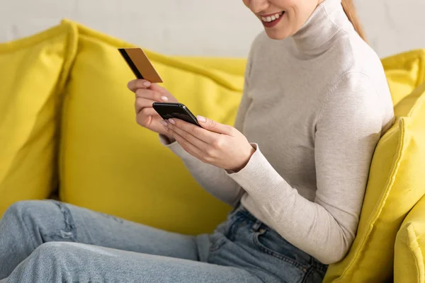Обрізаний вид усміхненої дівчини за допомогою смартфона і тримає кредитну картку на дивані — стокове фото
