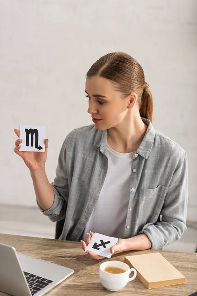 Attractive astrologer holding cards with zodiac signs beside book, laptop and coffee on table — Stock Photo