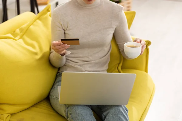Обрізаний вид жінки з кредитною карткою і ноутбуком п'є каву на дивані — стокове фото