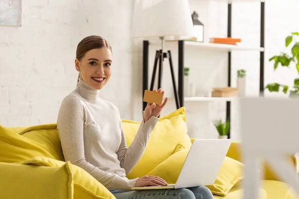 Hermosa chica que muestra la tarjeta de crédito mientras se utiliza el ordenador portátil y sonriendo a la cámara en el sofá - foto de stock