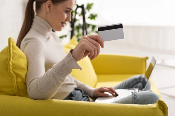 Focus selettivo della ragazza sorridente che mostra la carta di credito e utilizza il computer portatile sul divano — Foto stock