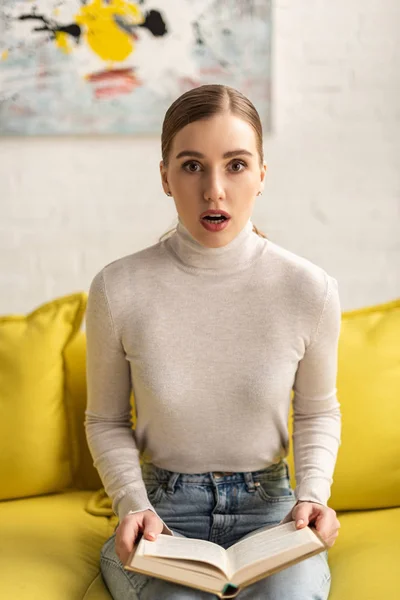 Choqué jeune femme regardant la caméra et tenant le livre sur le canapé — Photo de stock