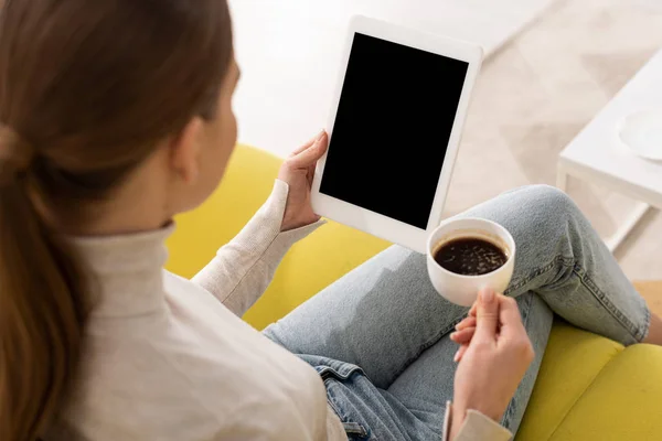 Vista sobre el hombro de la niña sosteniendo la tableta digital con pantalla en blanco y beber café en el sofá - foto de stock