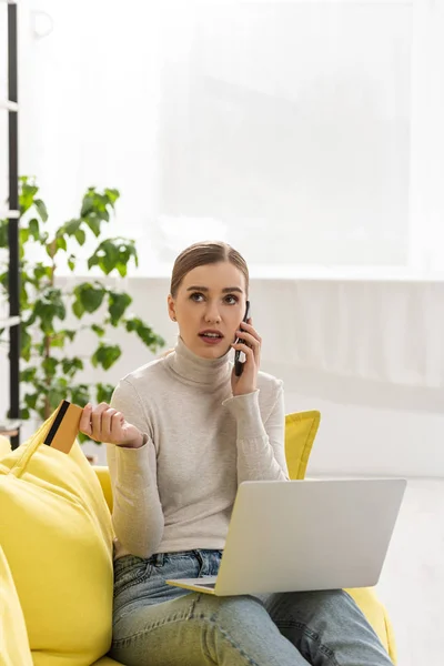 Mujer pensativa hablando en el teléfono inteligente mientras sostiene la tarjeta de crédito y el ordenador portátil en el sofá - foto de stock