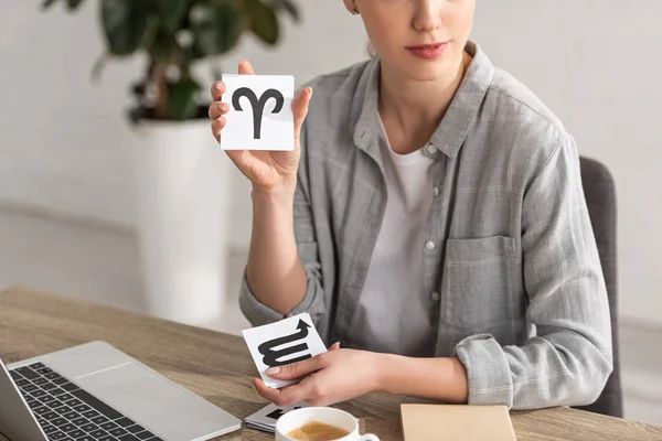 Cropped view of astrologer showing cards with zodiac signs beside coffee, book and laptop on table — Stock Photo