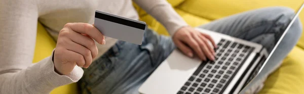 Cropped view of girl showing credit card and using laptop on sofa, panoramic shot — Stock Photo