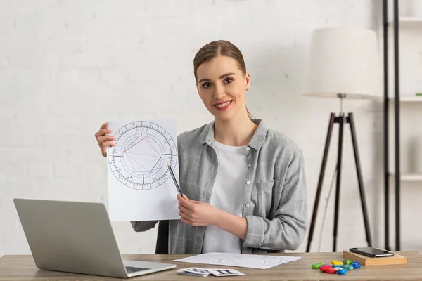 Schöne Astrologin zeigt Geburtshoroskop und lächelt in die Kamera — Stockfoto