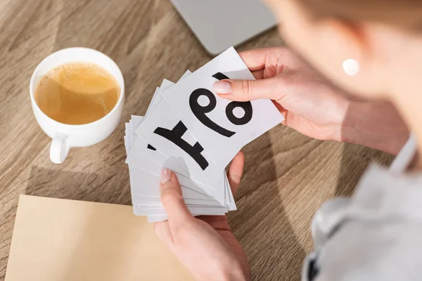 Cropped view of woman holding cards with zodiac signs beside coffee and laptop on table — Stock Photo