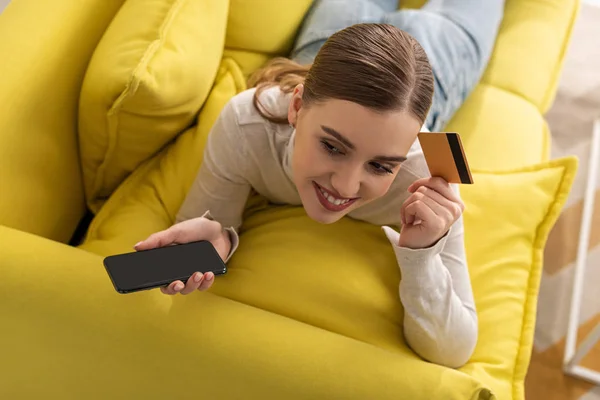 Vista aerea della donna sorridente che tiene carta di credito e smartphone mentre è sdraiata sul divano — Foto stock