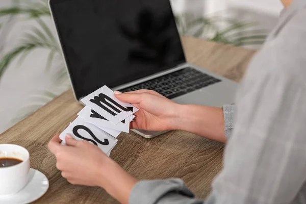 Cropped view of astrologer holding cards with zodiac signs beside laptop and coffee on table — Stock Photo