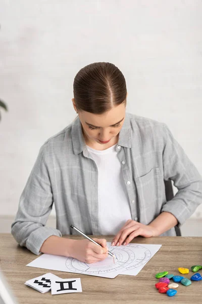Astrologer drawing natal chart beside cards and stones with zodiac signs on table — Stock Photo