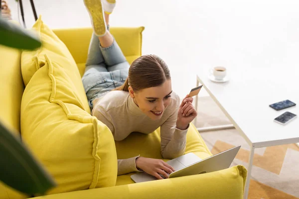 Smiling woman holding credit card and using laptop on sofa in living room — Stock Photo