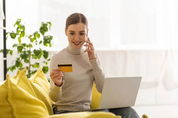 Femme souriante tenant un ordinateur portable et une carte de crédit tout en parlant sur un smartphone sur le canapé — Photo de stock